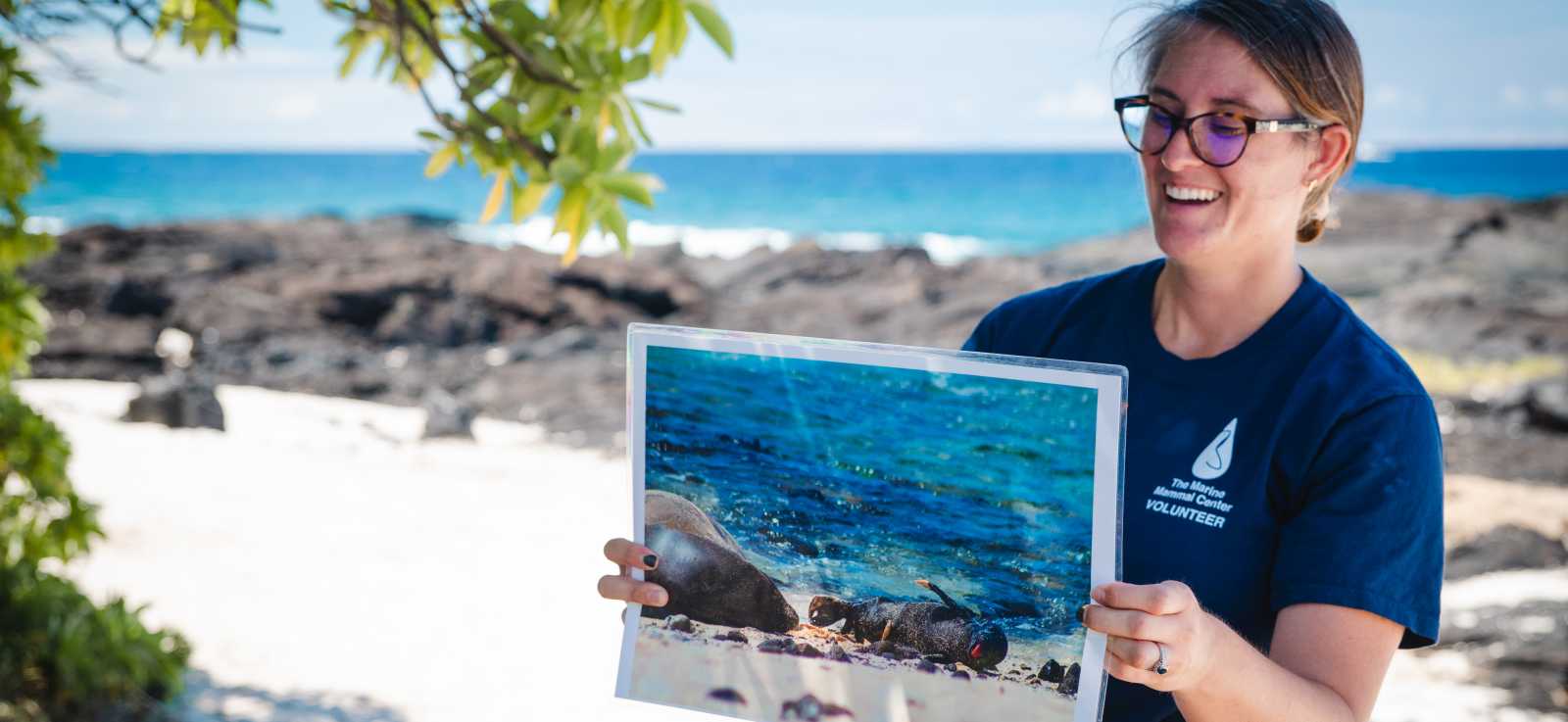 an educator from The Marine Mammal Center holds up an image of a baby monk seal while on the beach