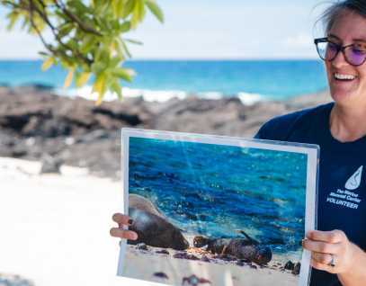 an educator from The Marine Mammal Center holds up an image of a baby monk seal while on the beach