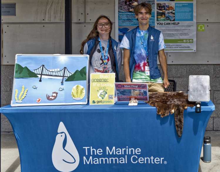 Two high school students stand behind a conservation education table with environmental posters and a sea otter pelt.