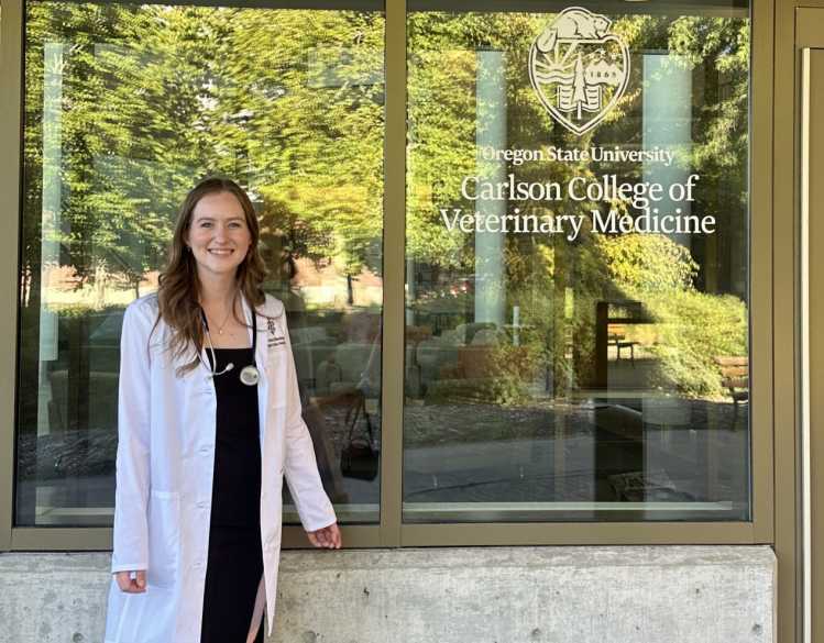 A veterinary student in a white coat stands in front of an Oregon State University Carlson College of Veterinary Medicine building.