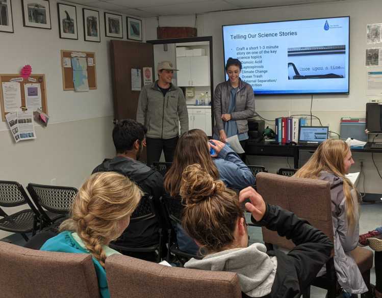 A student stands next to a screen that says “Telling Our Science Stories” while giving a presentation in front of a class.