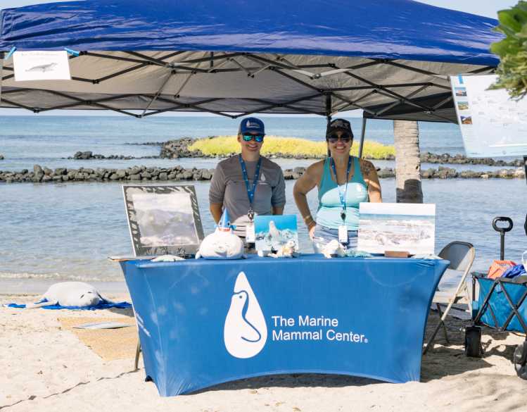 two staff members at a table on the beach with a blue tablecloth featuring The Marine Mammal Center's logo