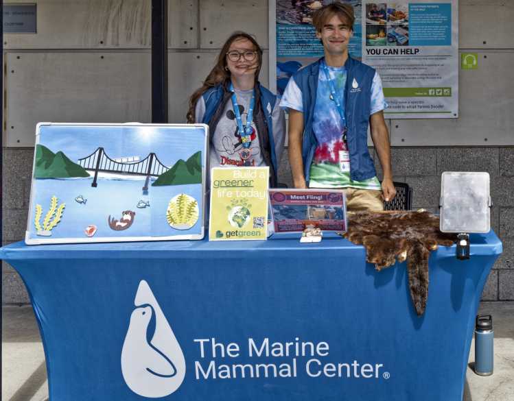 Two high school students stand behind an education table with environmental posters and a sea otter pelt.